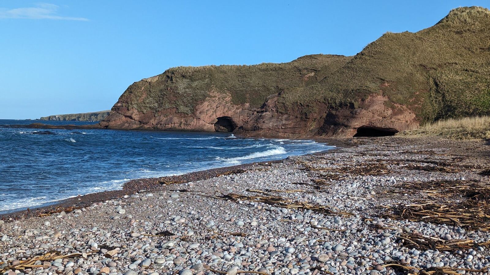 The secluded curving pebble beach of Aberdour Bay in Aberdeenshire, green cliffs and blue Moray Firth.