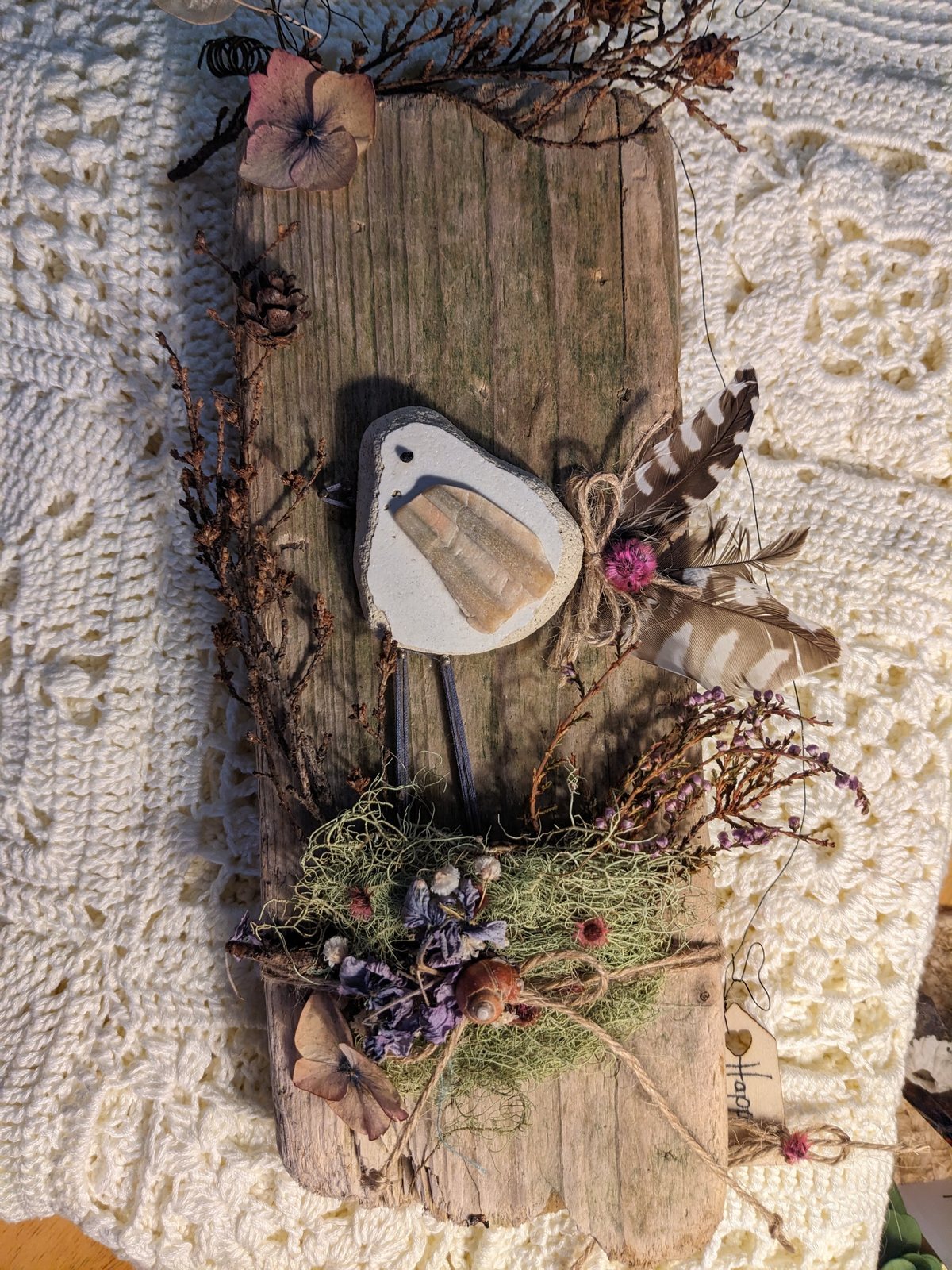 A handmade ceramic bird mounted on driftwood with feathers, dried heather and moss.