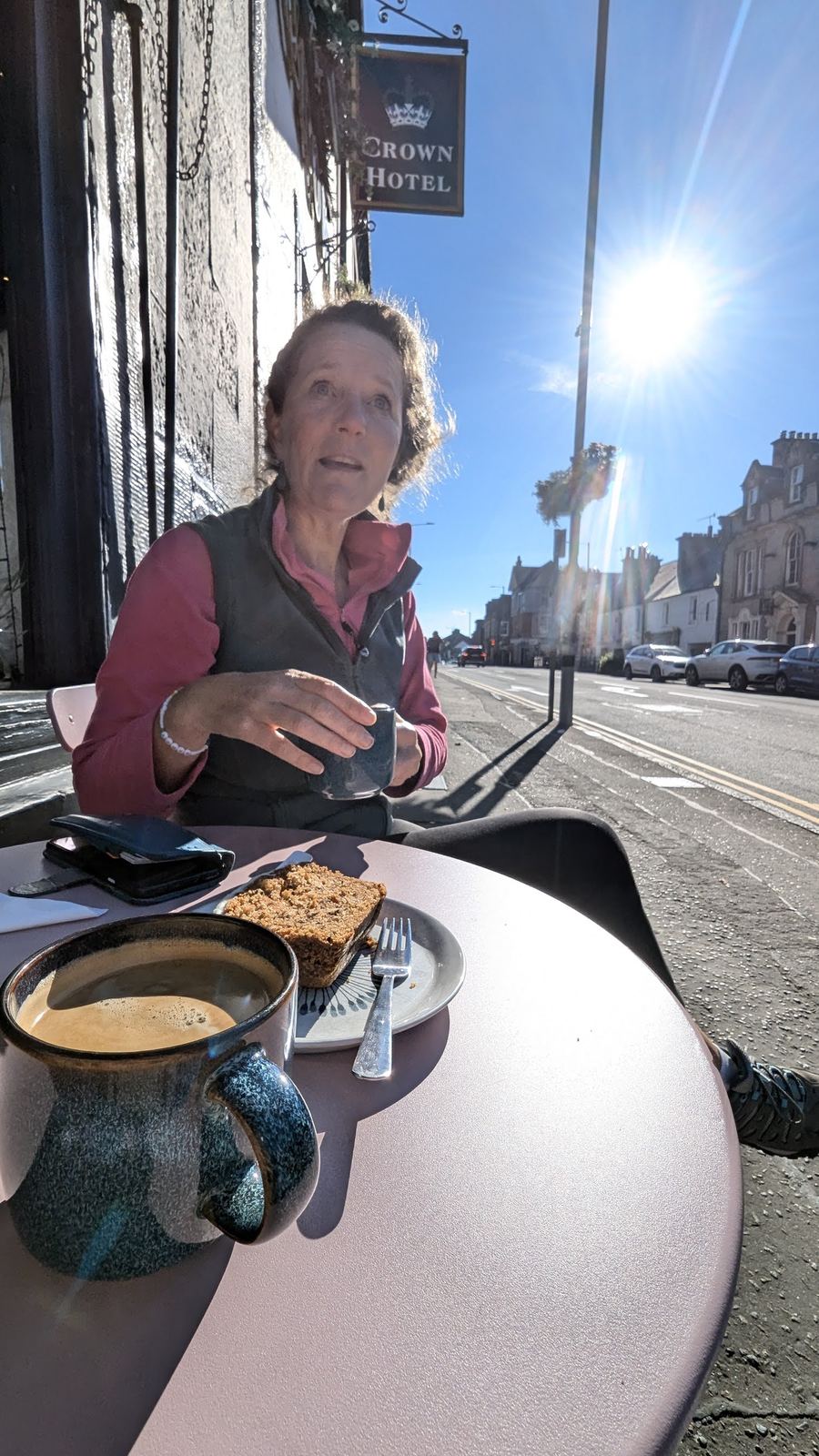 A sunny morning at an outdoor cafe table on Callander's wide Main Street, coffee and cake on the table, Victorian shopfronts and hills behind.