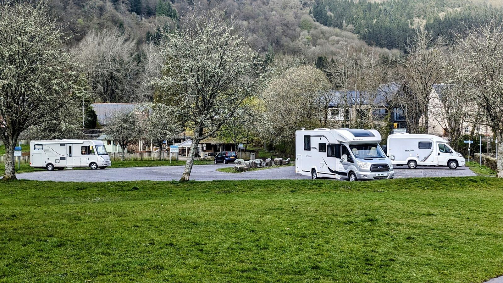 Motorhomes and campervans parked on a riverside area at Callander with spring blossom and hills behind.