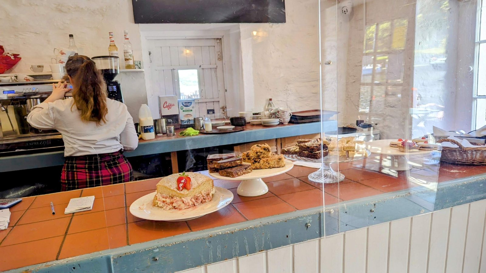 Inside the Crinan coffee shop, a server at the counter and cakes under a glass dome.
