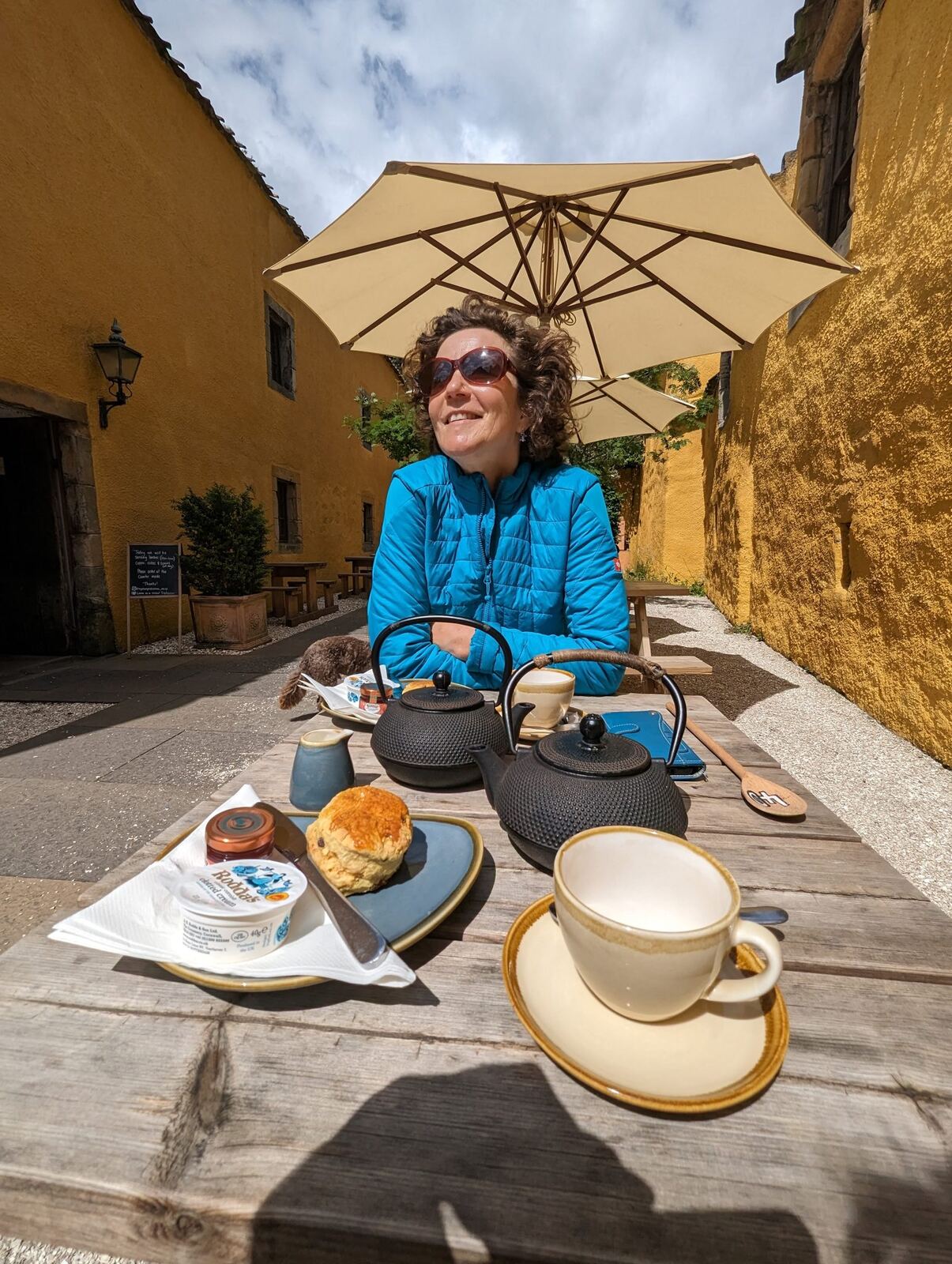A woman at a café table with cast-iron teapots and scones in a sunny Culross courtyard.