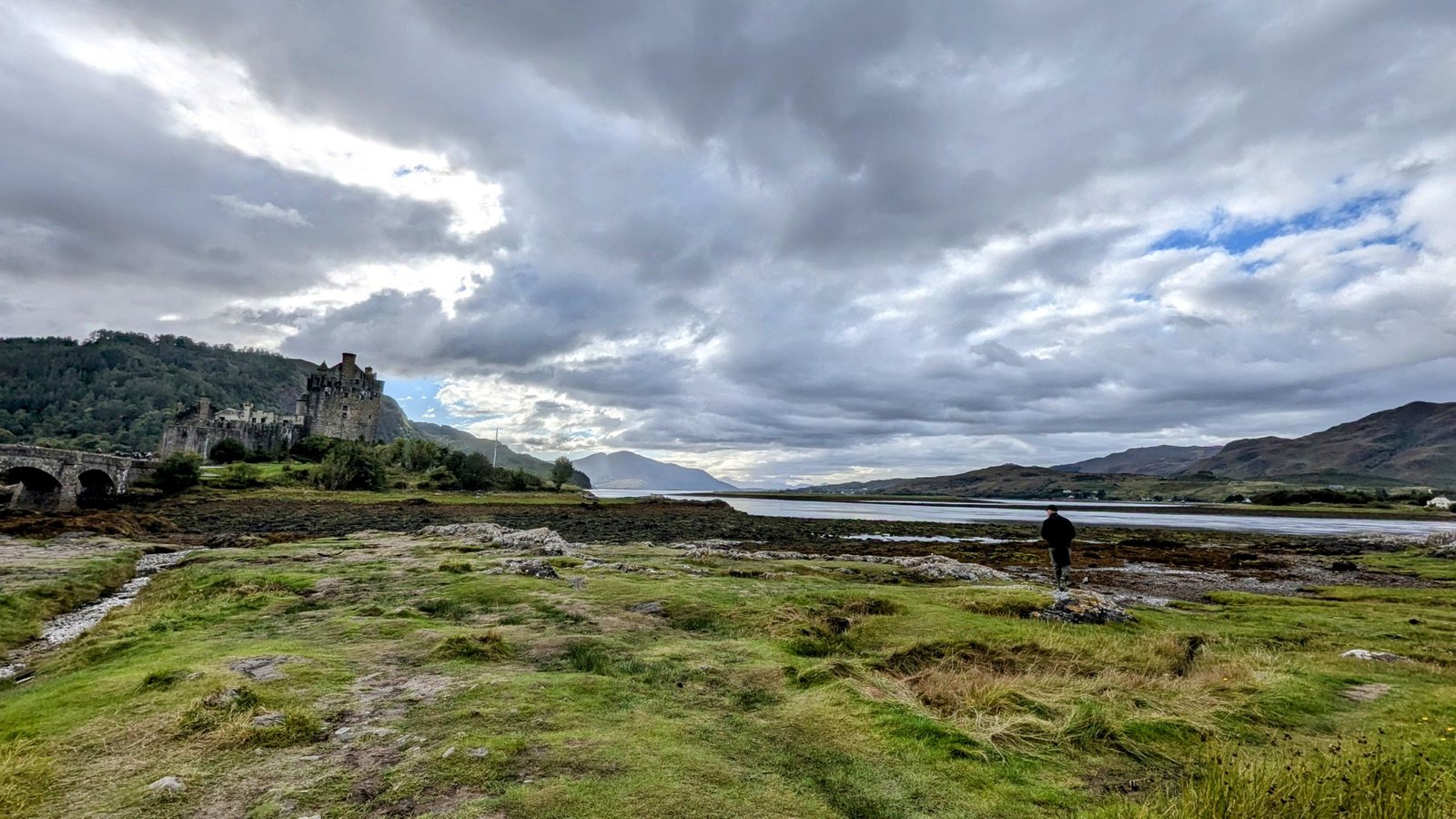 The castle and its arched bridge from the grassy foreshore, a walker and dog on the path, hills of Kintail beyond