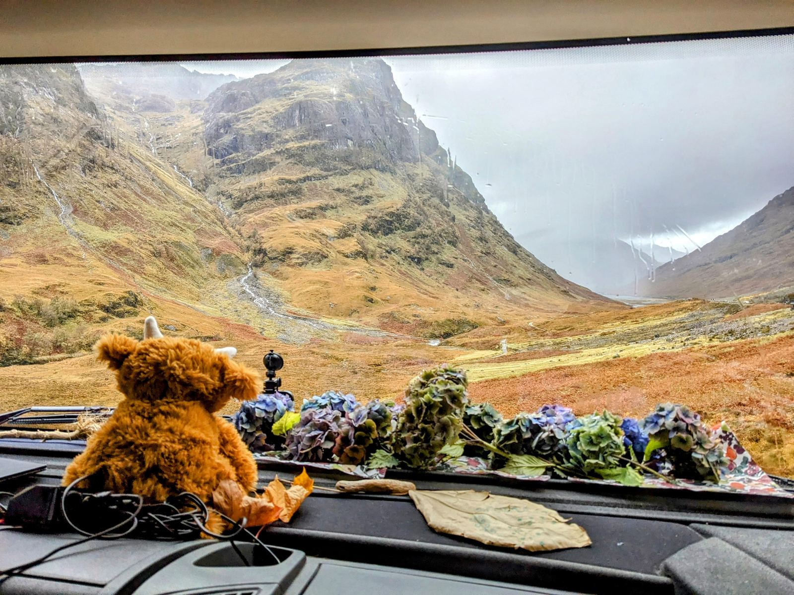 View from the cab approaching a deep mountain glen, steep peaks rising ahead, toy Highland cow on the dashboard.