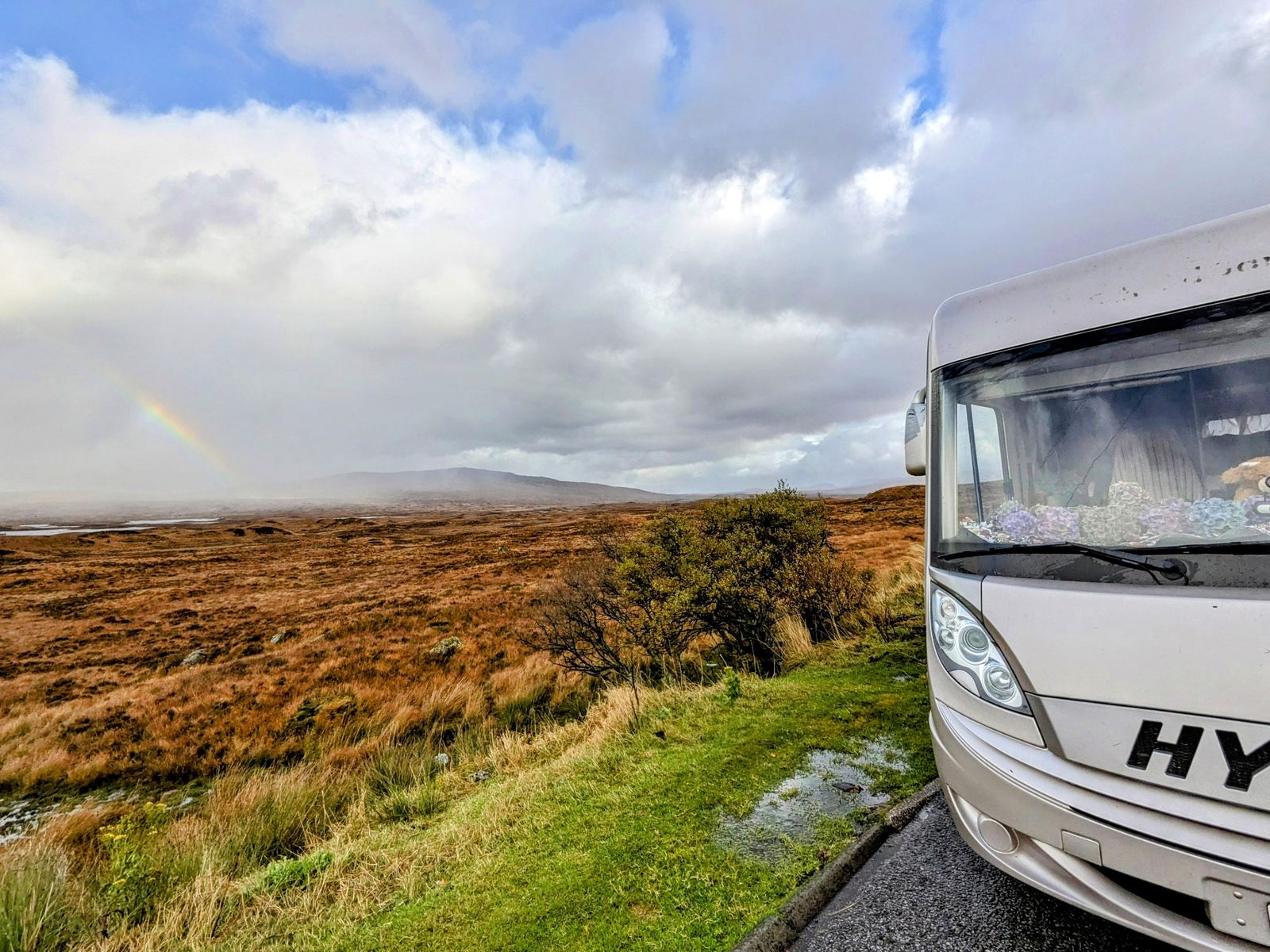 A Hymer motorhome parked on the empty moor road across Rannoch Moor with a faint rainbow over the golden bog.