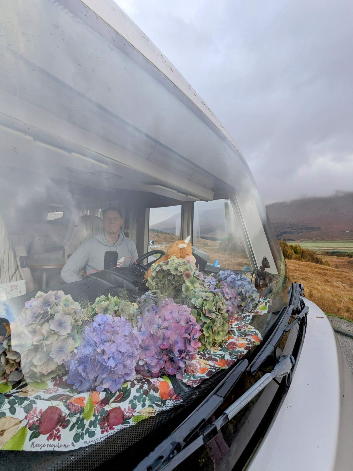 View from a motorhome cab crossing the empty A82 over Rannoch Moor in autumn, low cloud on the hills, flowers and a toy Highland cow on the dashboard.