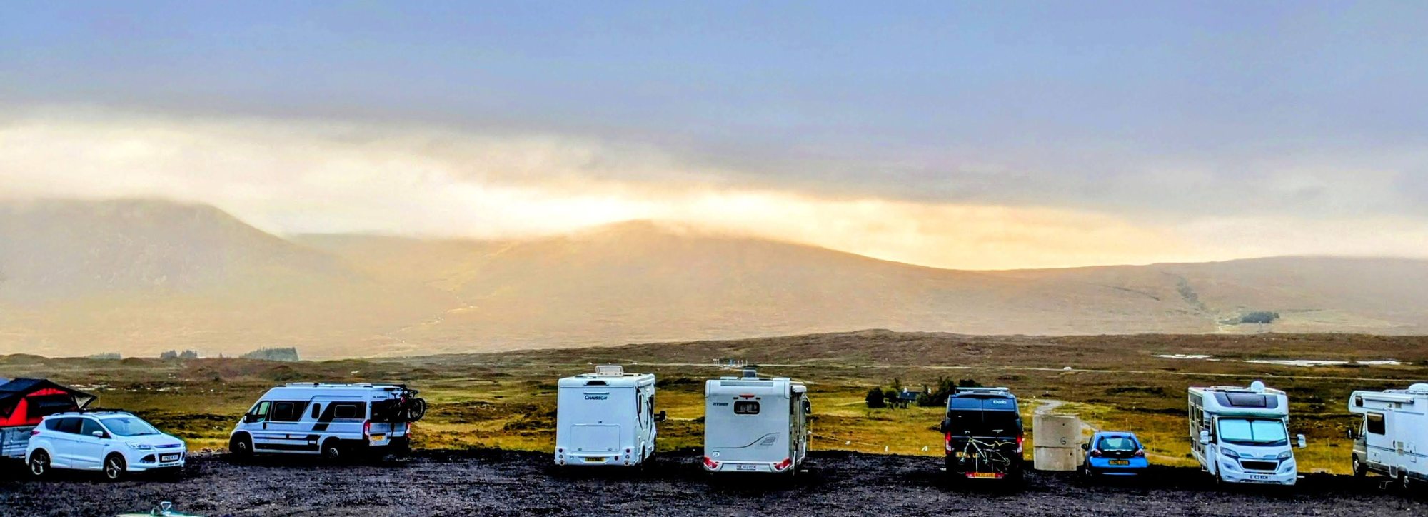 Motorhomes and campervans lined up at a moorland car park above Glen Coe as golden light breaks over the hills.