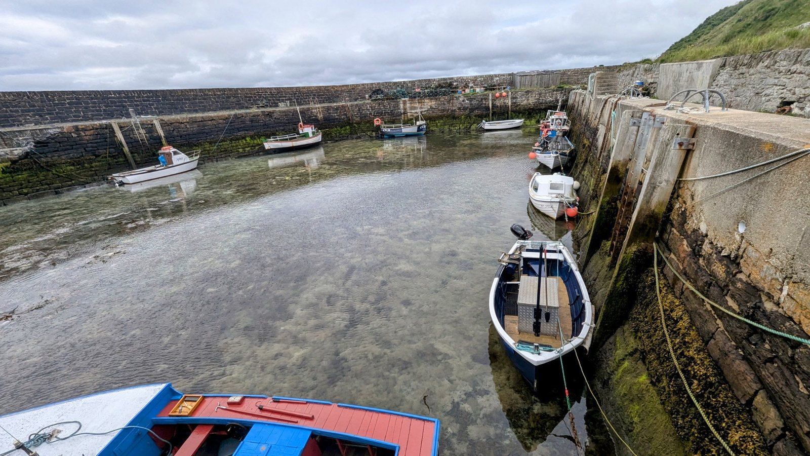 Wide view of Keiss Harbour at low tide with several small fishing boats resting in shallow clear water, ringed by old stone harbour walls and a green hillside.