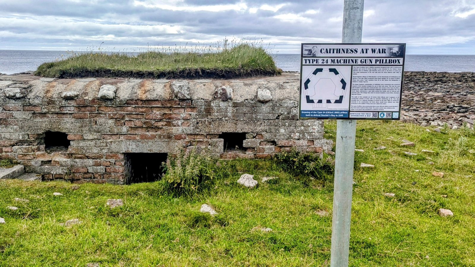 A grass-topped WWII Type 24 hexagonal brick pillbox beside a 'Caithness at War' interpretation board on the shore at Keiss, sea behind.
