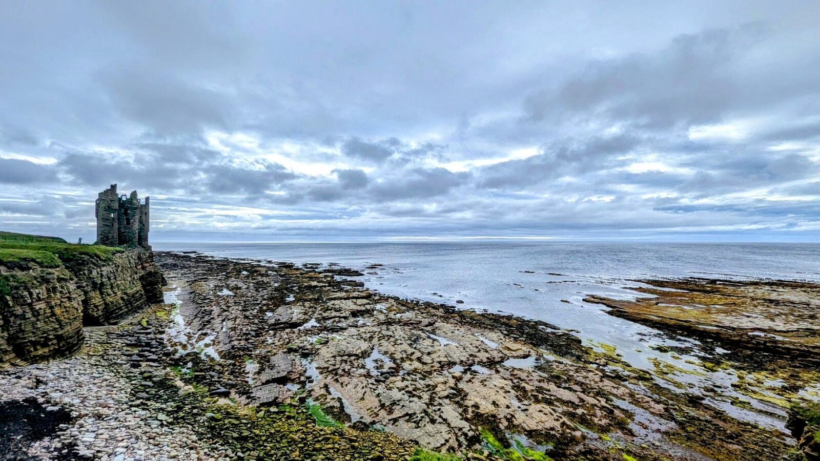 Wide dawn view of the Caithness coast at Keiss with the castle ruin on the left headland and rock platforms stretching into a calm sea.