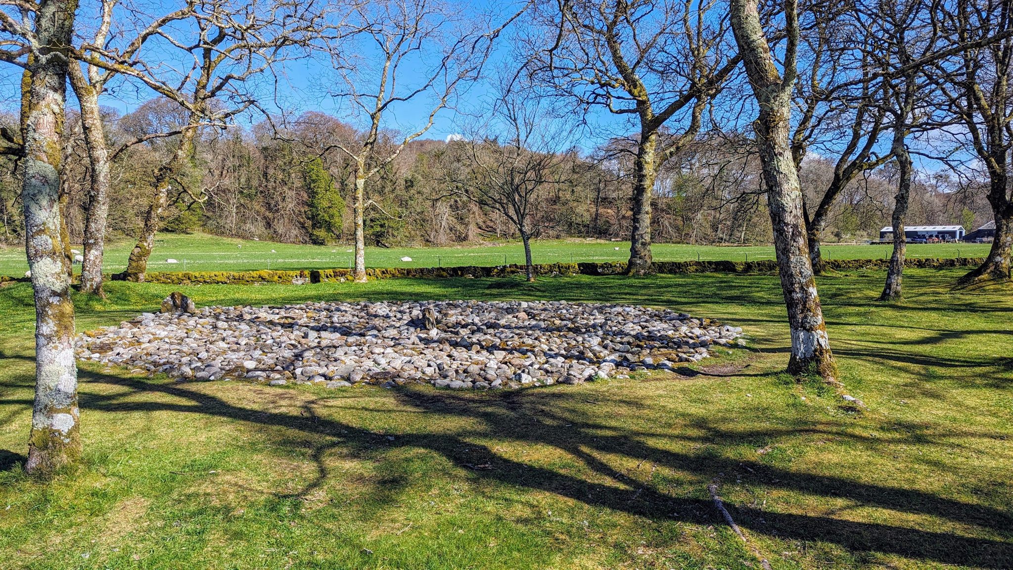 A large round Bronze Age burial cairn of grey stones set among trees in parkland at Kilmartin Glen.
