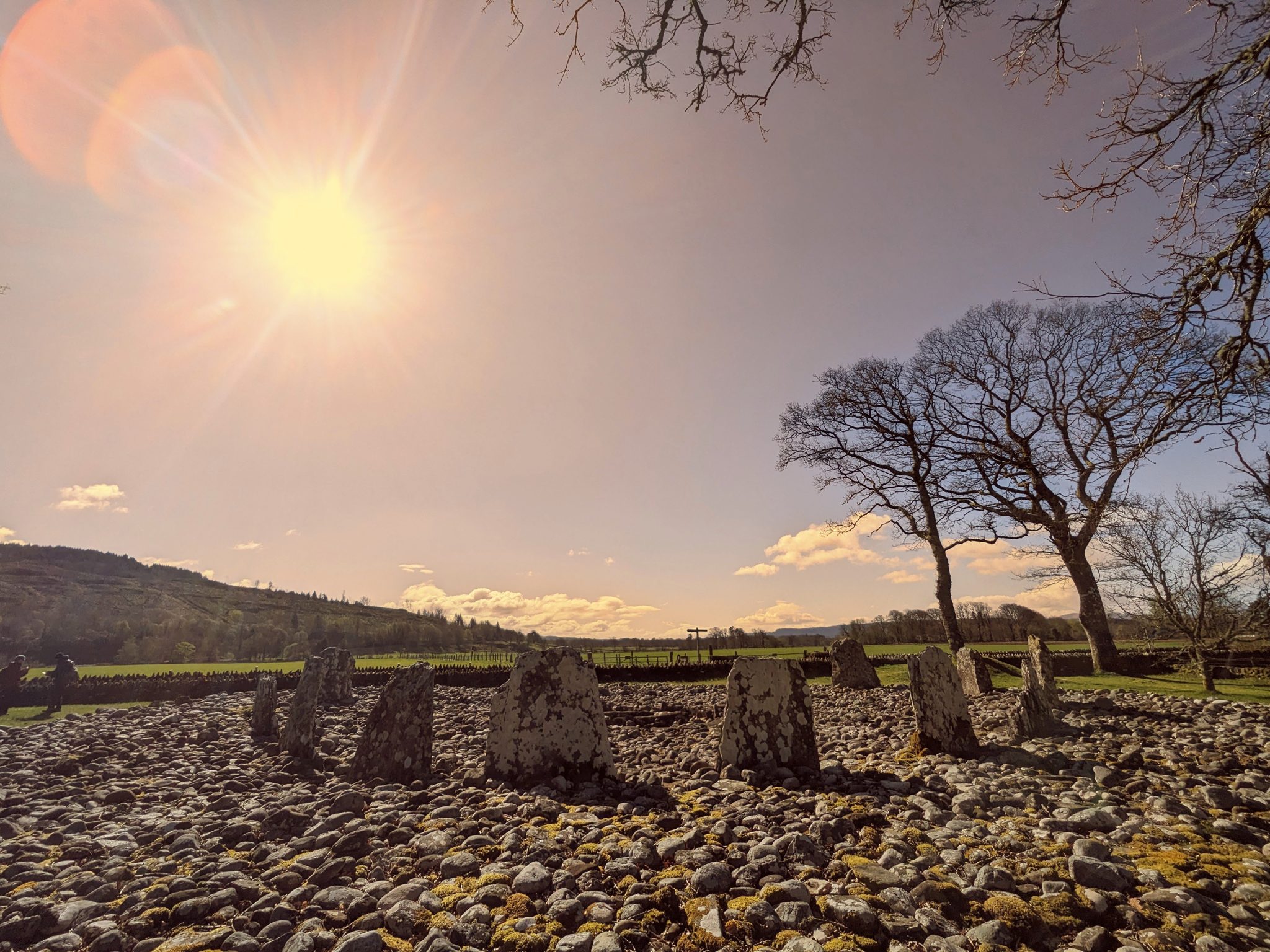 Standing stones and a Bronze Age cairn at Kilmartin Glen silhouetted against a low sun bursting through the sky.