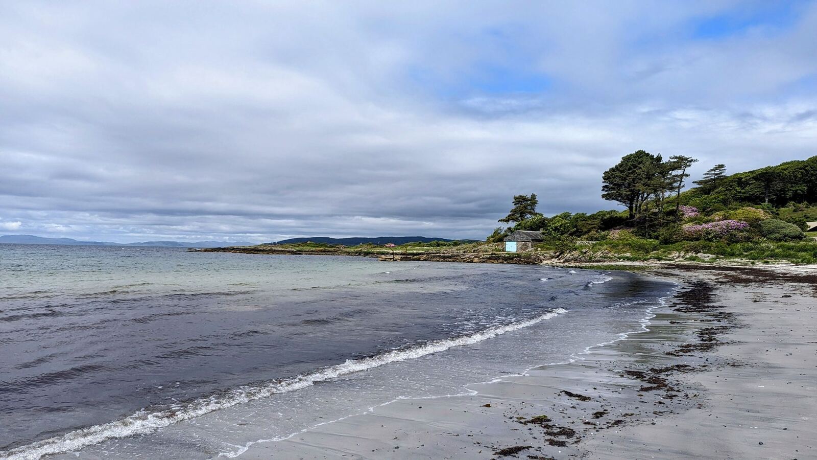 The curve of the pebble beach on the Kintyre Way with a wooded point and islet beyond.