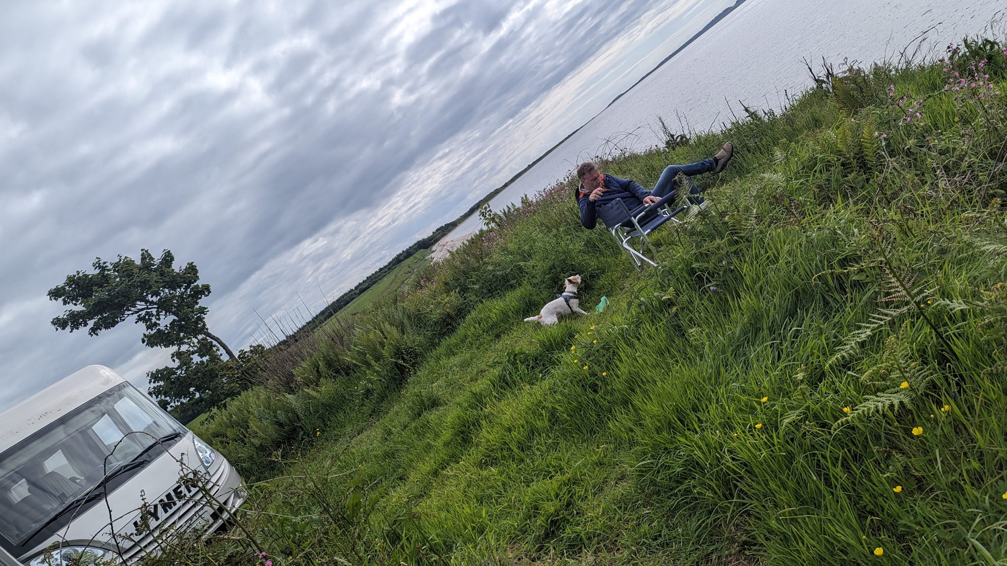 A woman relaxing in a camping chair on the grass by the motorhome with Lillie the dog, looking out to sea on the Kintyre coast.
