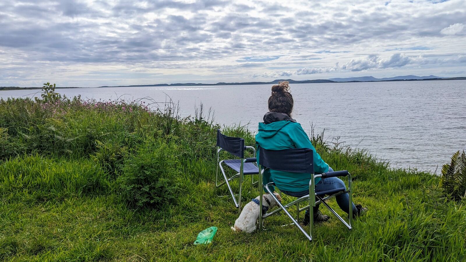 A woman sitting in a camping chair on the grassy bank above the sea, looking out to Arran on the east Kintyre coast.
