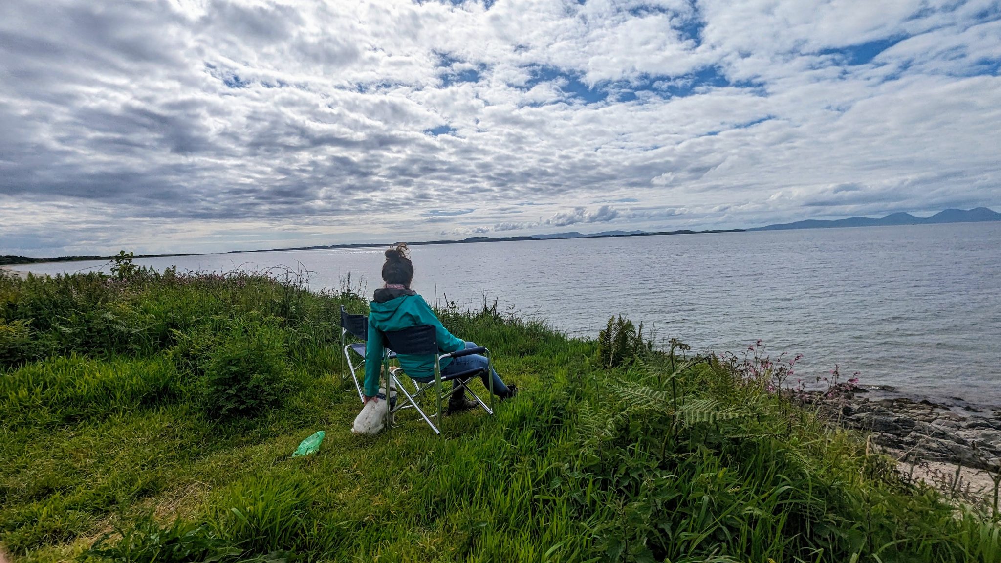 A woman sitting looking out across the Kilbrannan Sound to Arran from the grassy shore on the Kintyre Way.