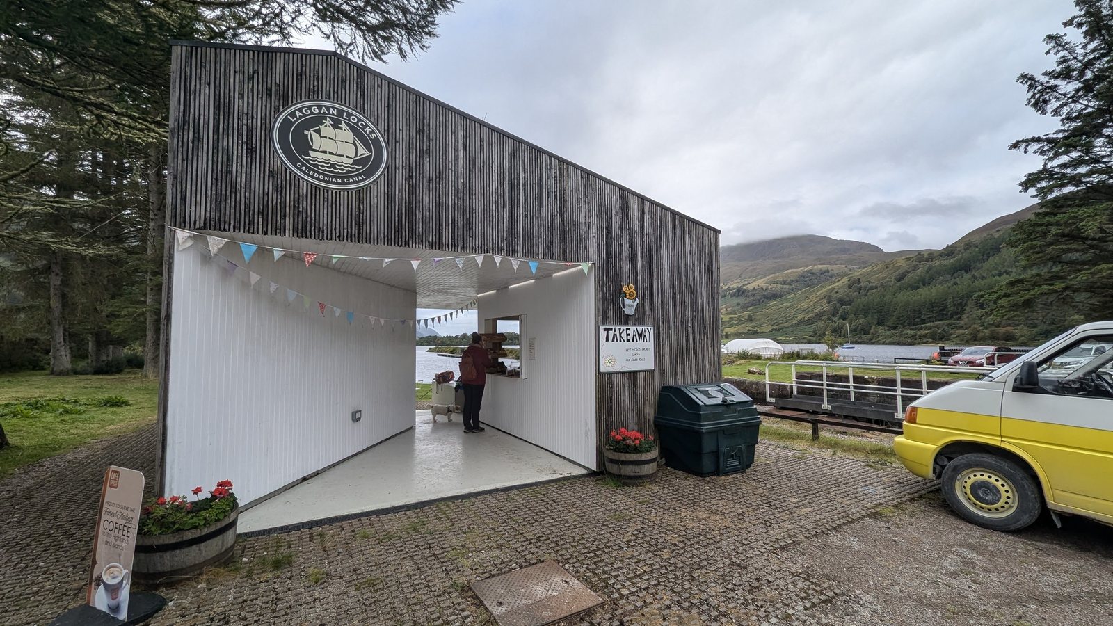 The Laggan Locks takeaway coffee kiosk on the Caledonian Canal, bunting and flower barrels, with a yellow VW T4 campervan parked outside and Lillie at the serving window.