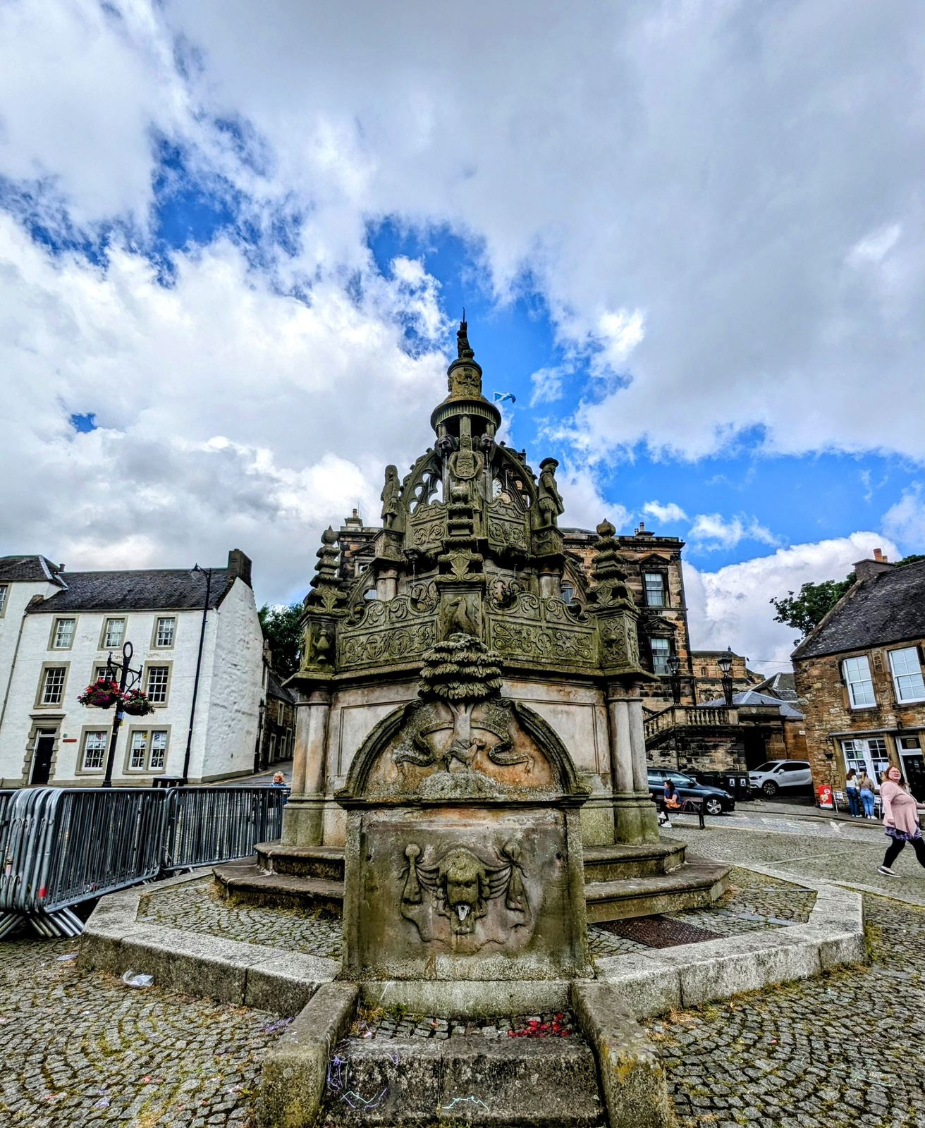 The ornately carved stone Cross Well fountain on the High Street at Linlithgow under a blue sky.