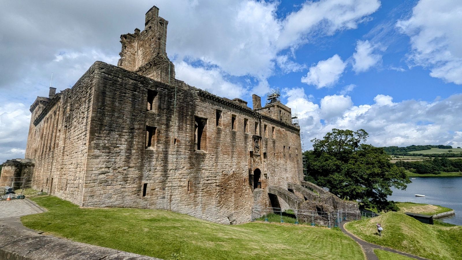 The tall ruined stone walls of Linlithgow Palace above green lawns beside the loch under a big sky.