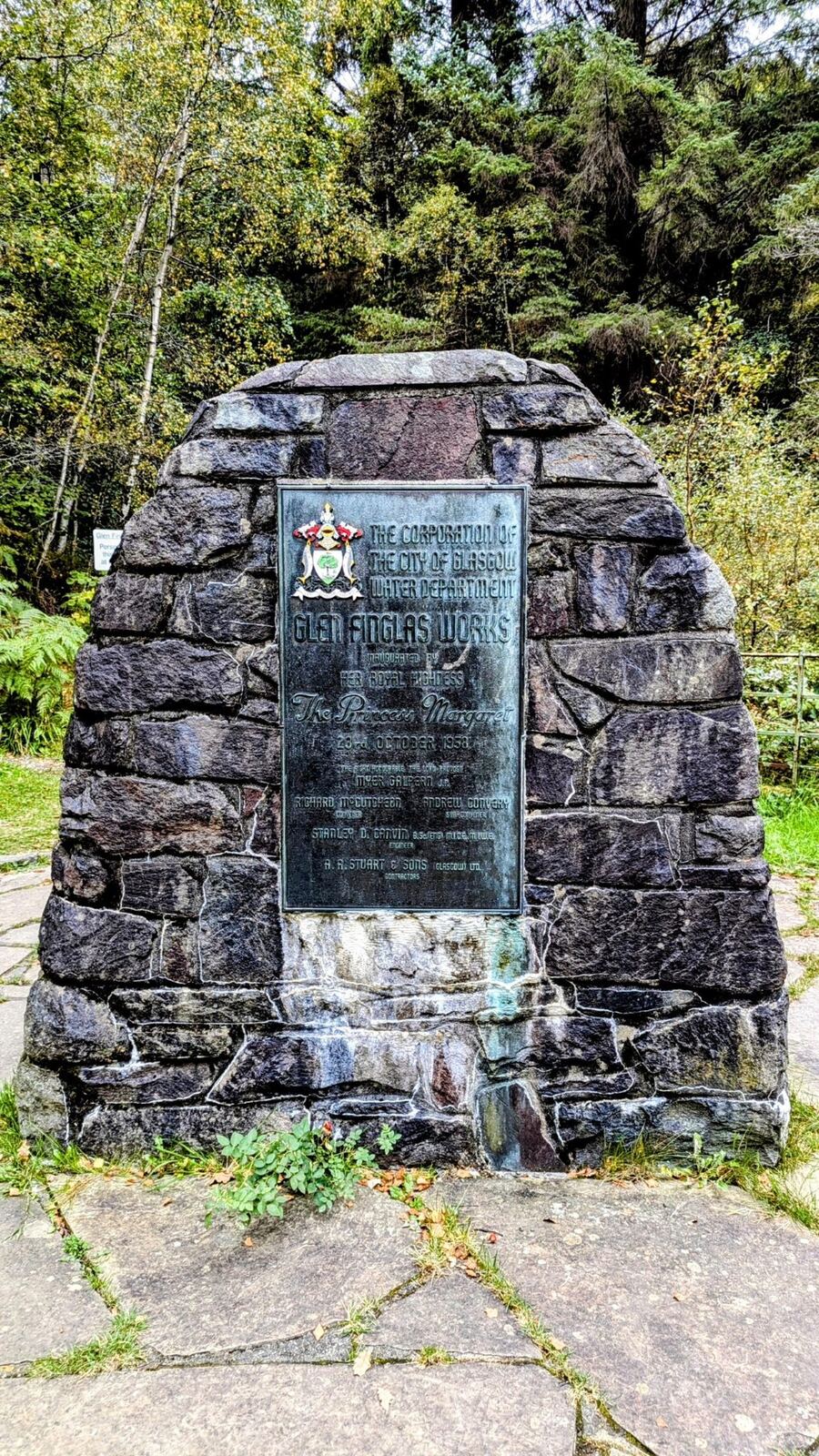 A stone memorial cairn with an inscribed plaque beside a path in the Trossachs near Loch Katrine.