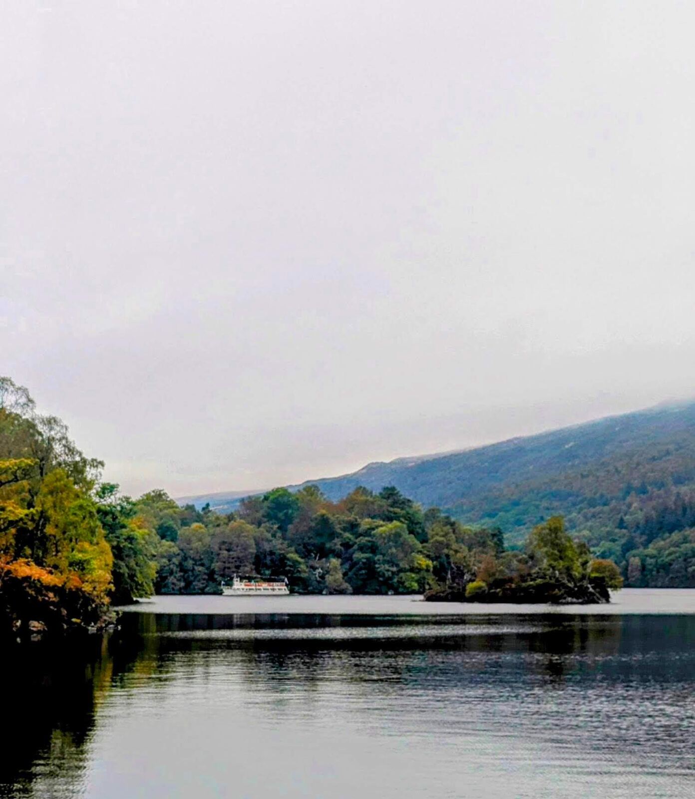 A misty, still morning on Loch Katrine, the wooded shore and hills fading into low cloud over calm water.