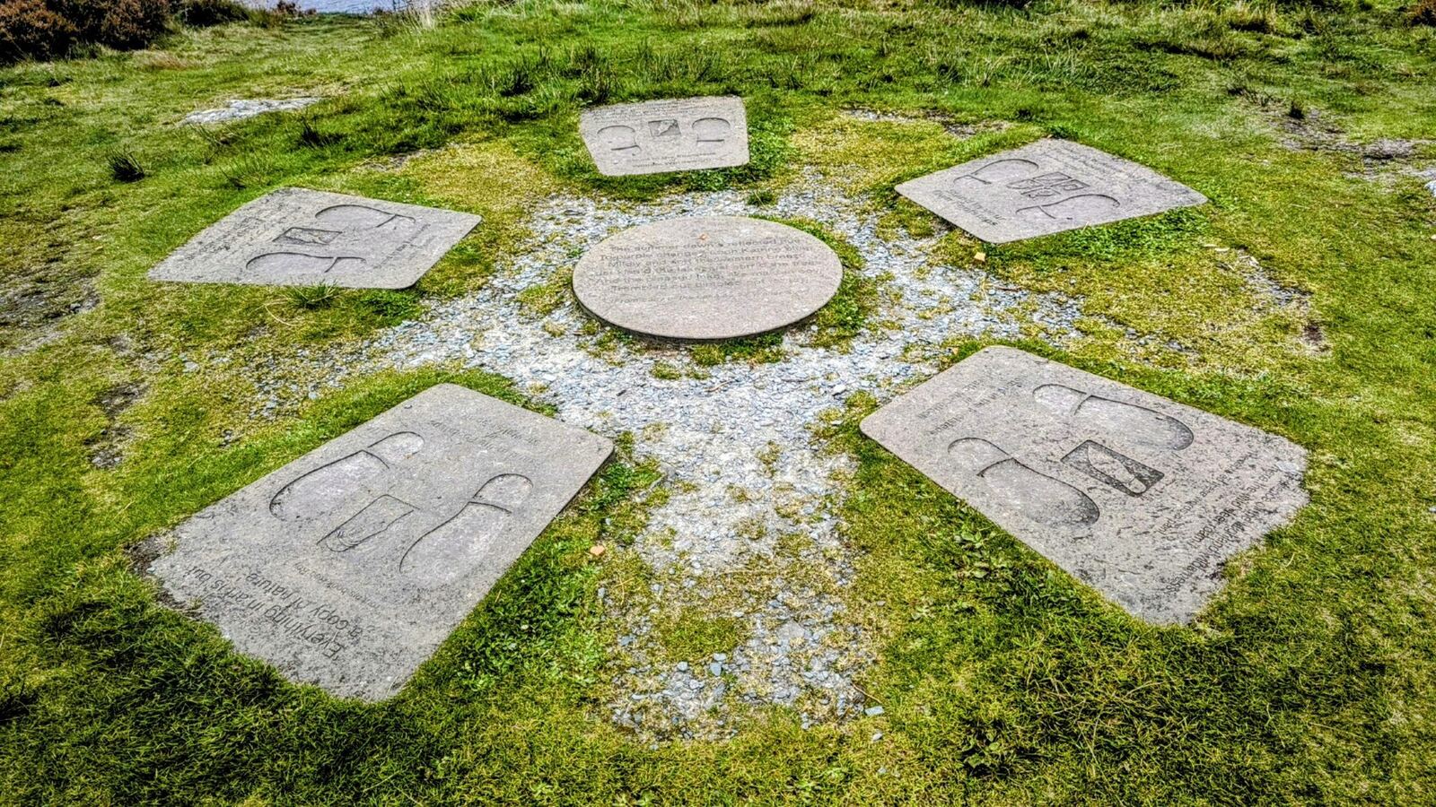 An engraved circle of stone slabs set in the grass near Loch Katrine, a central round stone carved with a poem.