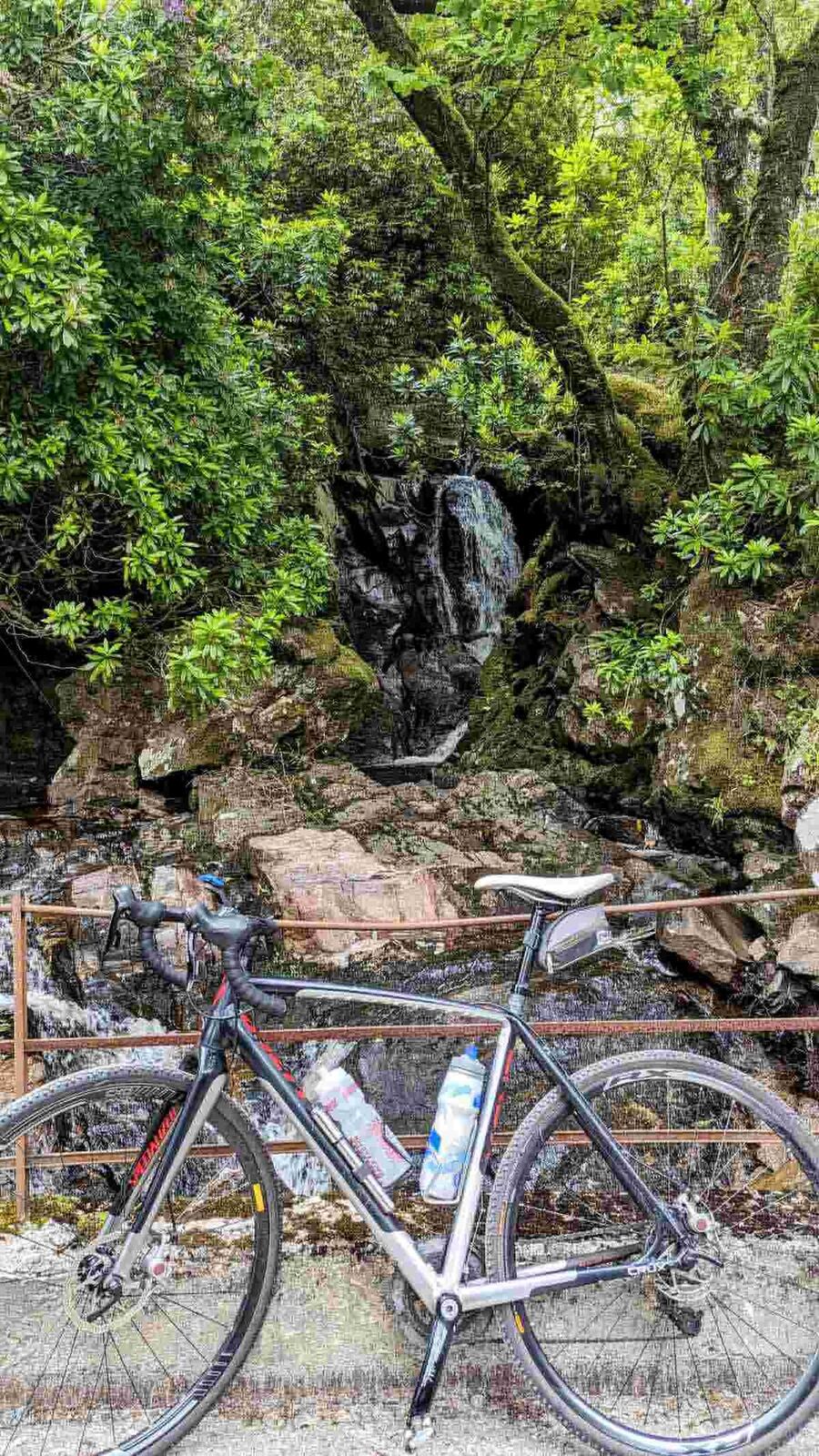 A gravel bike leaning by a mossy waterfall on the shore path of Loch Riddon.