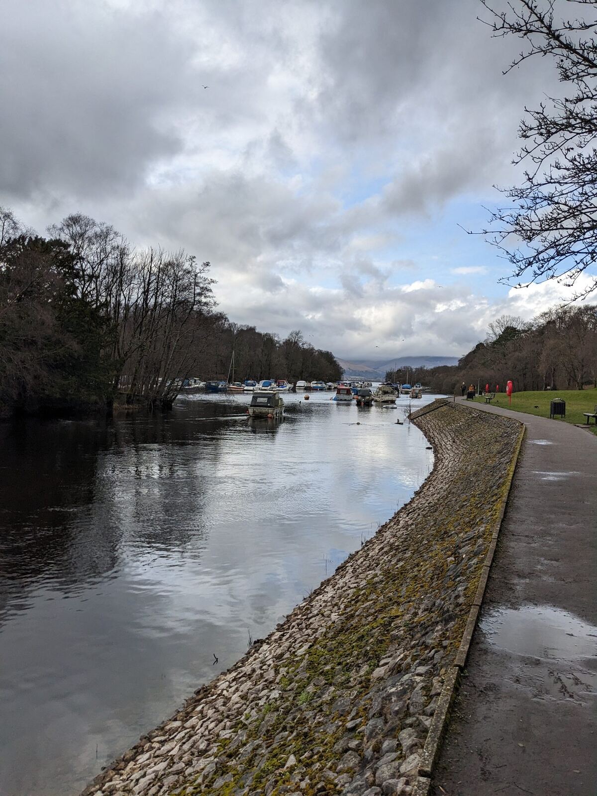 The River Leven at Balloch flowing out of Loch Lomond, a paved shore path curving away, boats and parkland alongside.