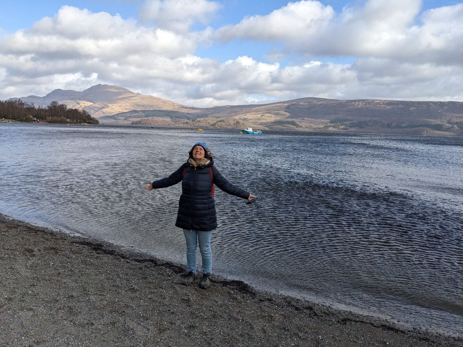 A woman standing arms-out on the pebbly shore of Loch Lomond, hills across the calm water.