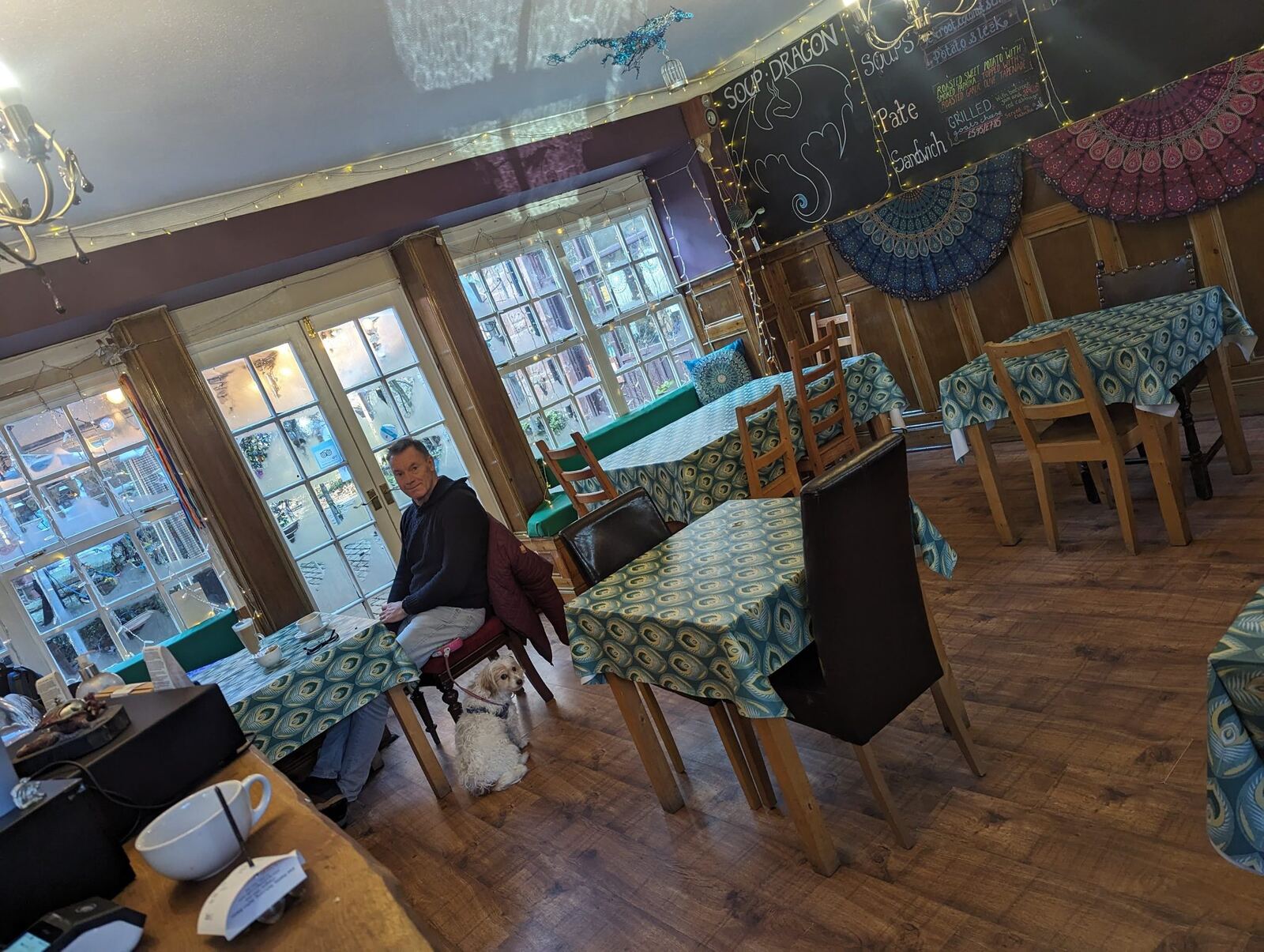 The interior of a village café at Balmaha with peacock-patterned tablecloths and a soup menu chalkboard, a man and a small dog at a table.