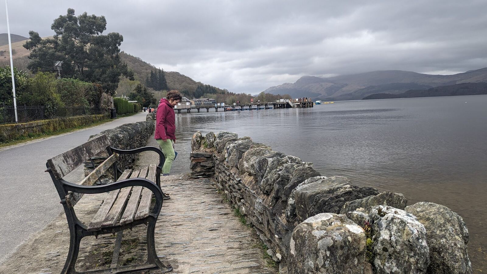 The Luss shore promenade on Loch Lomond, stone wall and benches, Ben Lomond and the hills beyond.