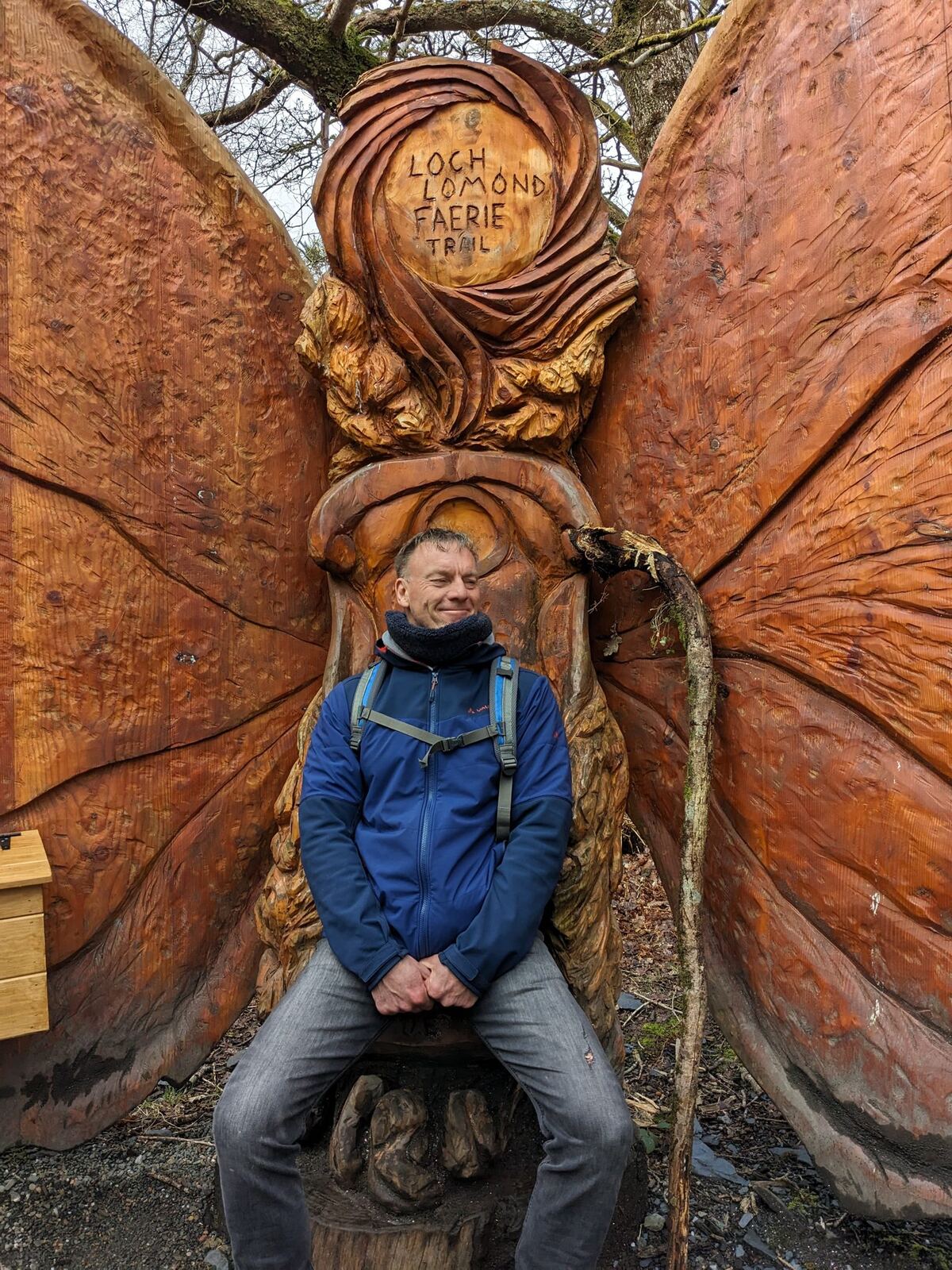 A man sitting in a large carved wooden winged throne on the Loch Lomond fairy trail at Luss.