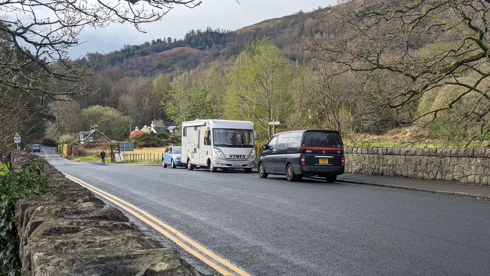 A motorhome and car parked on a quiet road by the loch near Luss, hills of Loch Lomond behind — the old free overnight bridge spot, now double-yellowed.