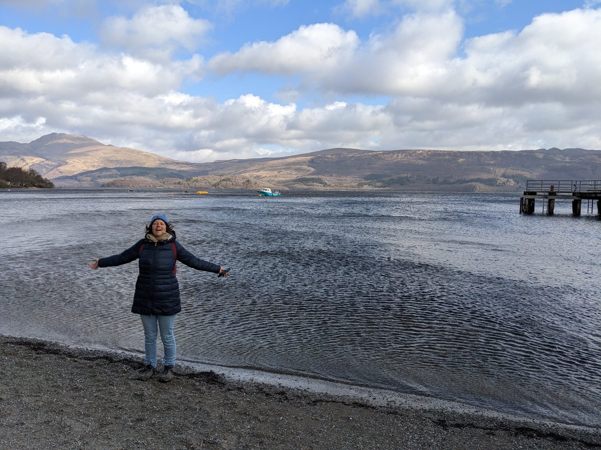 A visitor with arms wide on the shore of Loch Lomond at Luss, the loch and hills beyond.