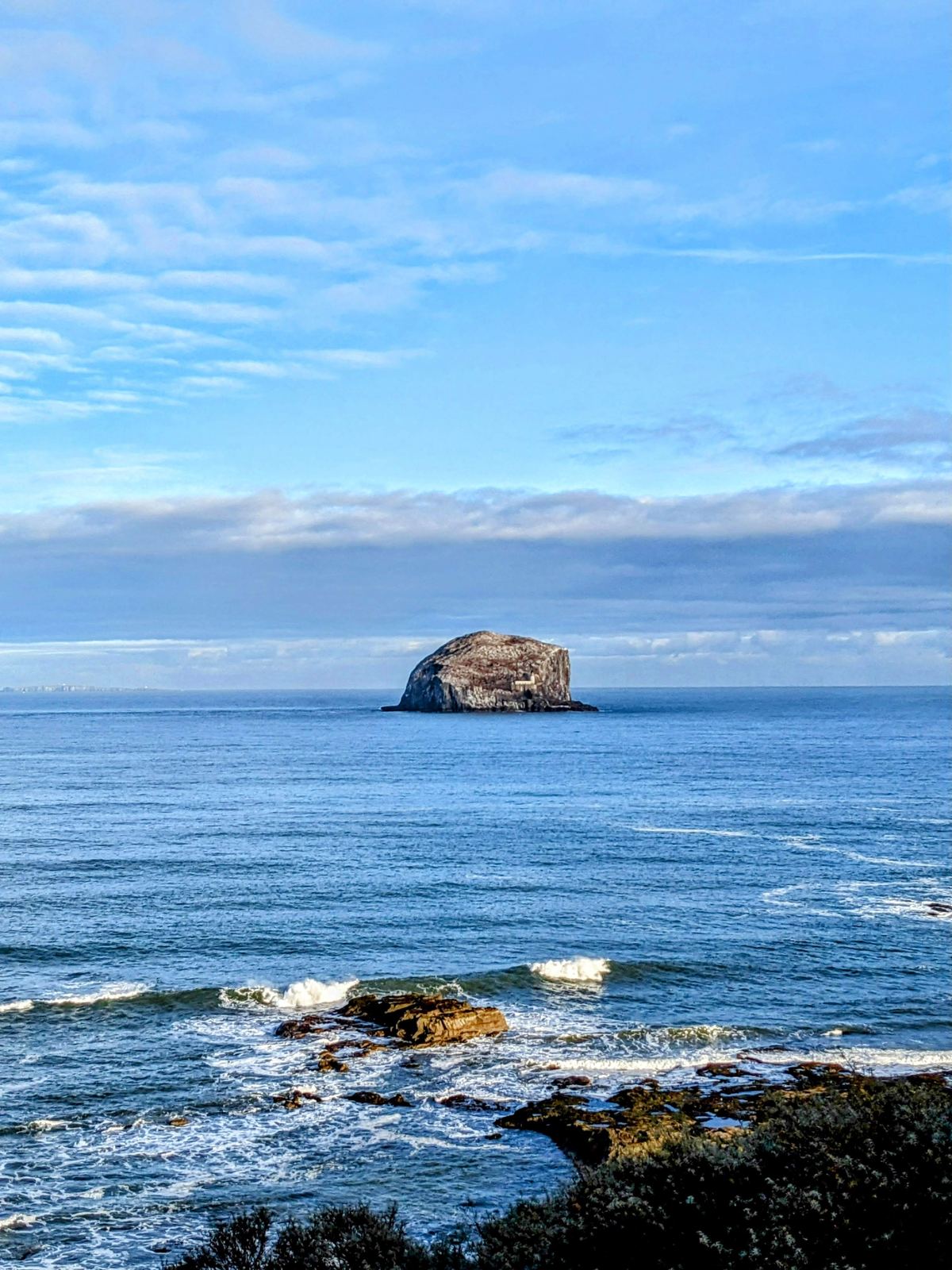 The white dome of the Bass Rock rising out of a choppy blue Firth of Forth, seen from the rocky shore at North Berwick.