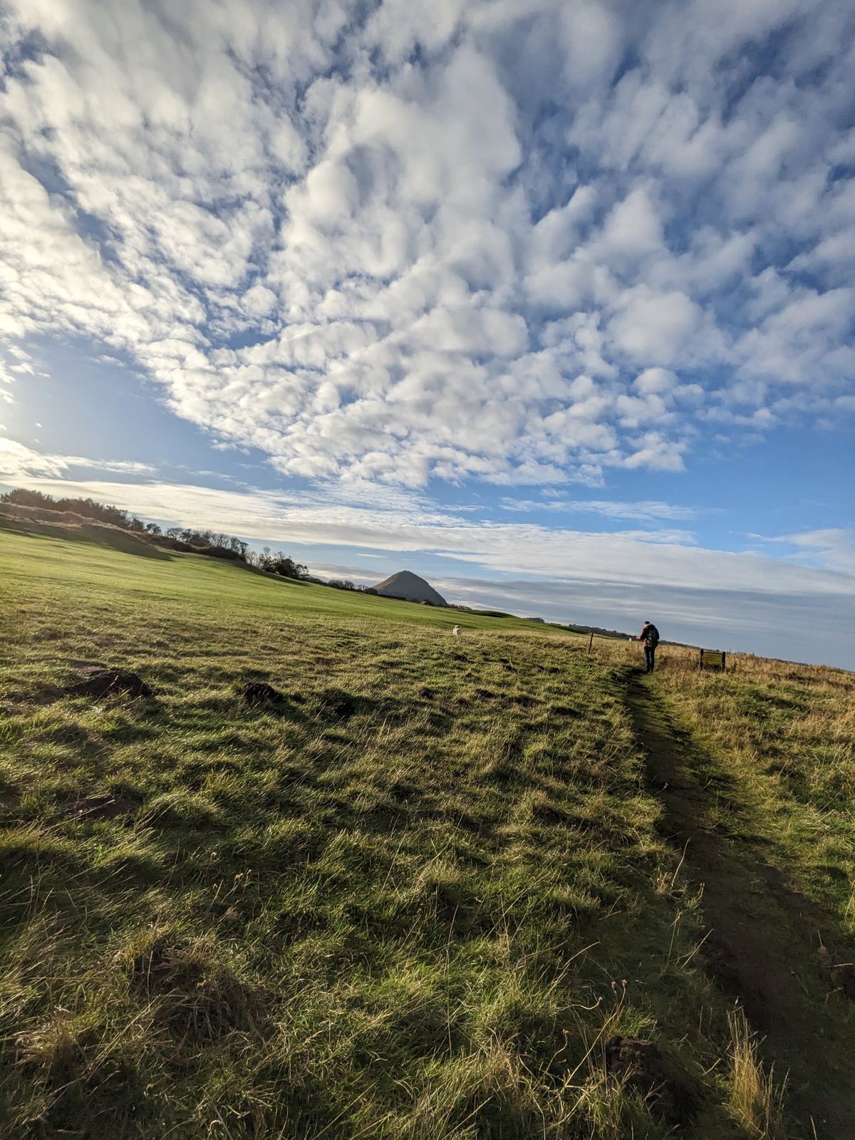 A walker on a grassy coastal path along the North Berwick shore under a huge cloud-streaked sky, islands out to sea.