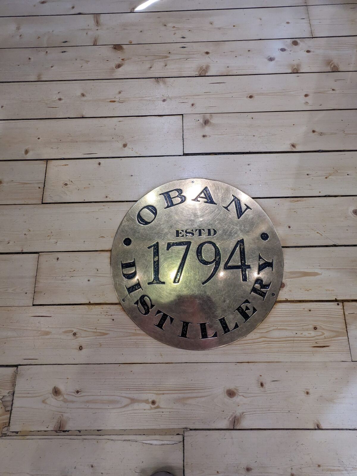 A brass 'Oban Distillery Estd 1794' plaque mounted on pale wood.