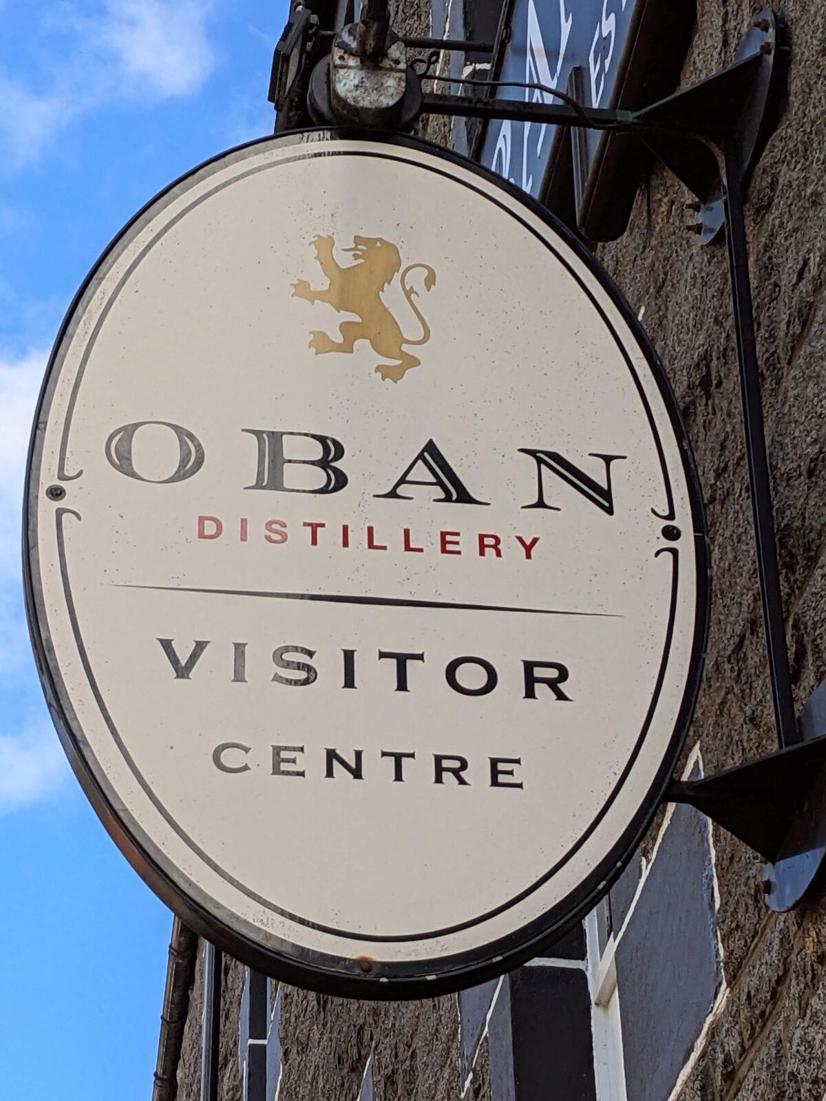 The round hanging sign of Oban Distillery Visitor Centre against a blue sky.