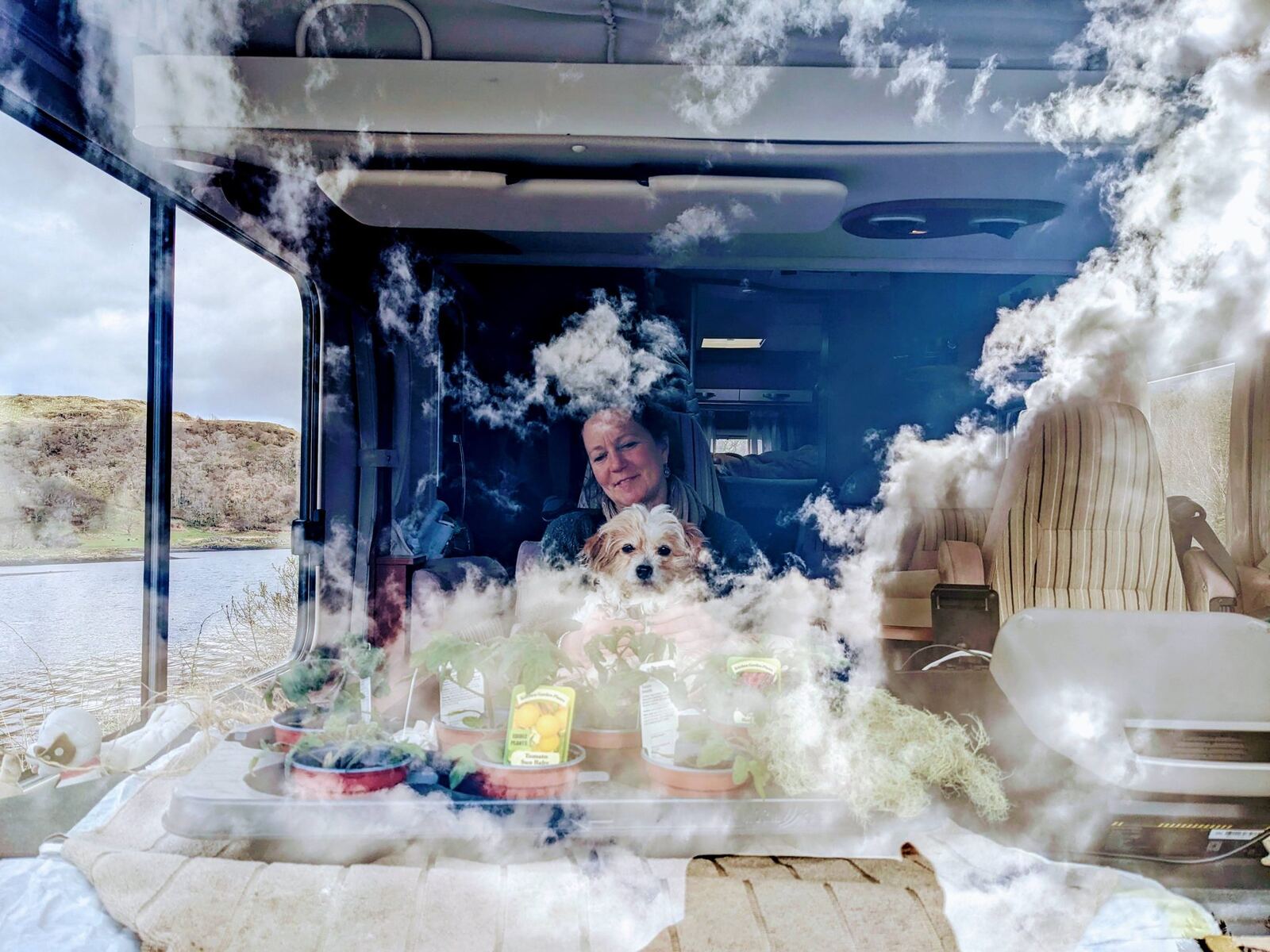 A woman and a small dog sitting inside a motorhome, sky and clouds reflected in the window, at a quiet spot south of Oban.