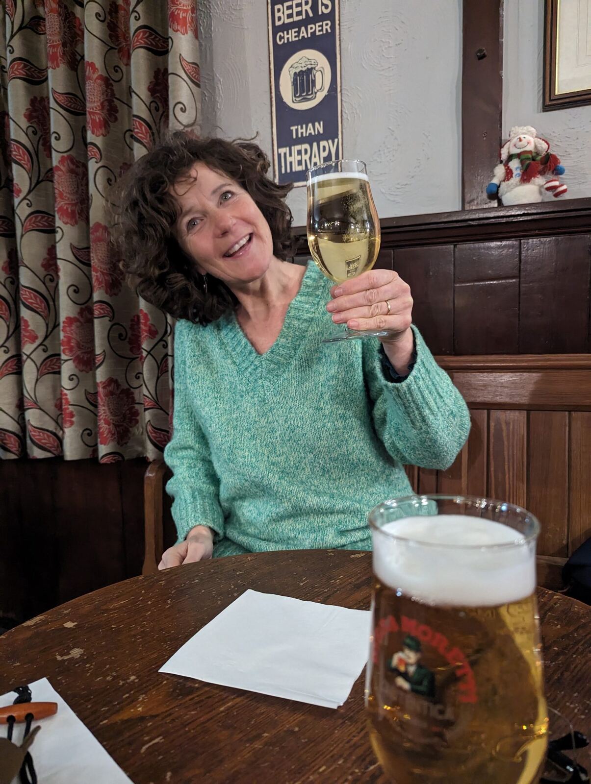 A woman in a green jumper smiling and holding up a pint of lager in the Flotterstone Inn near the Pentland Hills, a sign reading beer is cheaper than therapy behind her.
