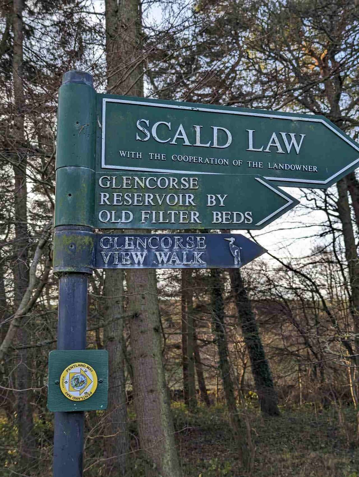 Green fingerpost signpost in the Pentland Hills reading Scald Law, Glencorse Reservoir by Old Filter Beds and Glencorse View Walk, with a Pentland Hills waymarker disc.