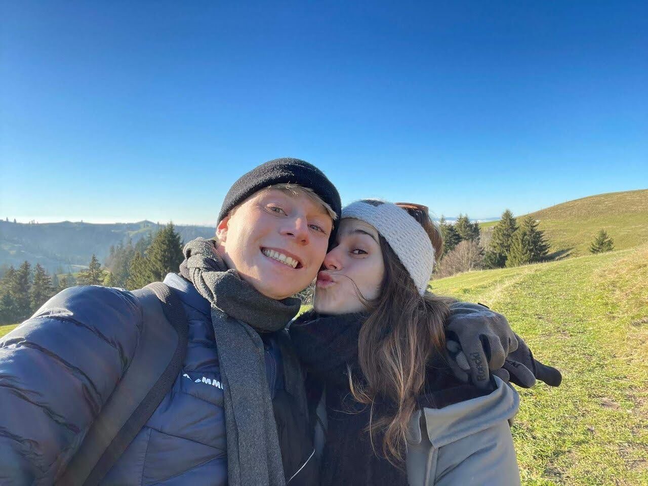 Two walkers in warm winter gear taking a selfie on a grassy Pentland hilltop under a clear blue winter sky.