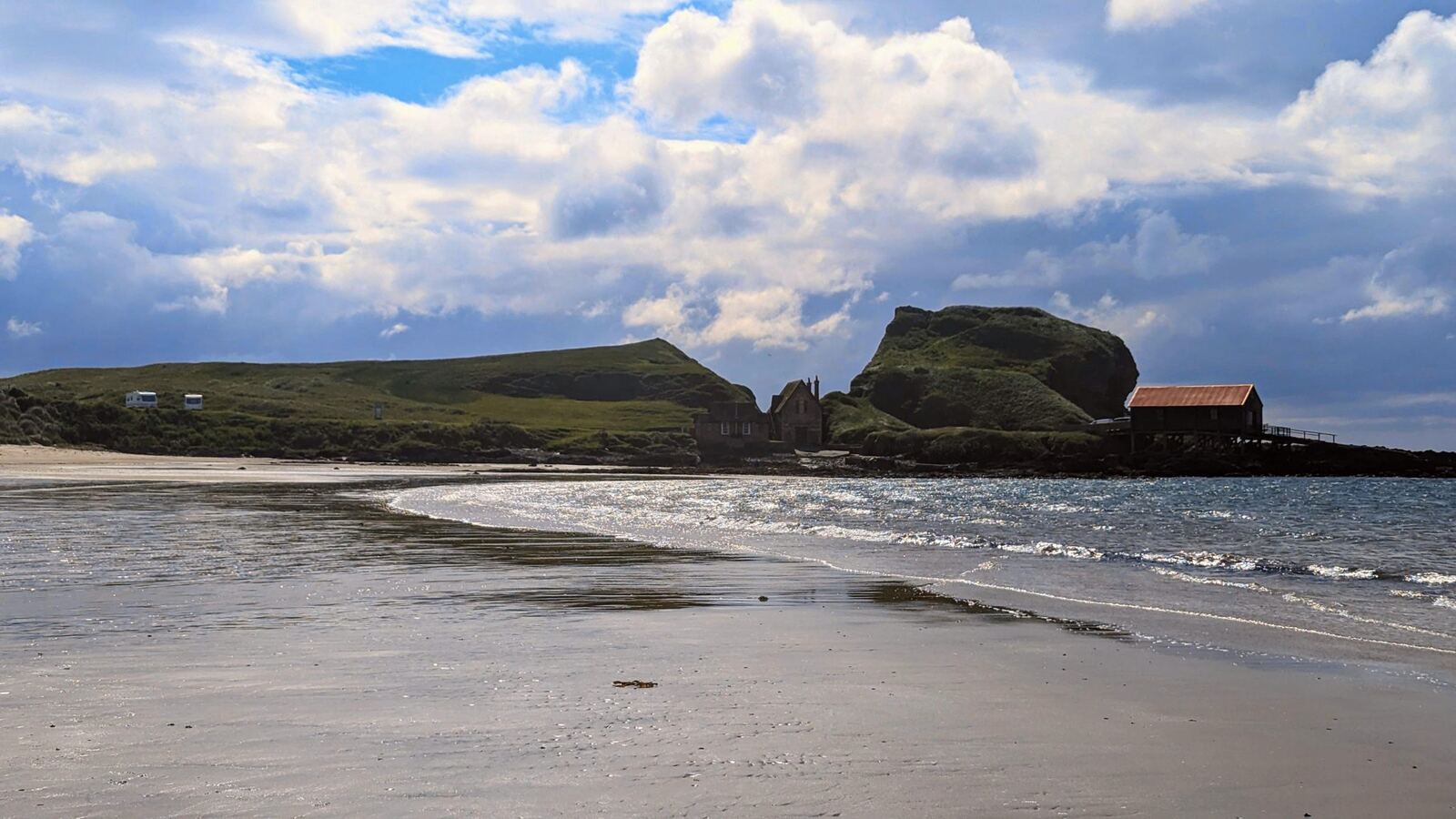 The wide sandy beach at Southend, Kintyre, with a rock stack and lifeboat-style building across the sand at low tide.