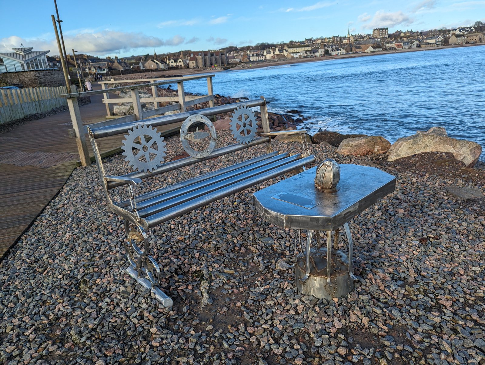 A metal bench and little wagon sculpture on the shingle at Stonehaven.