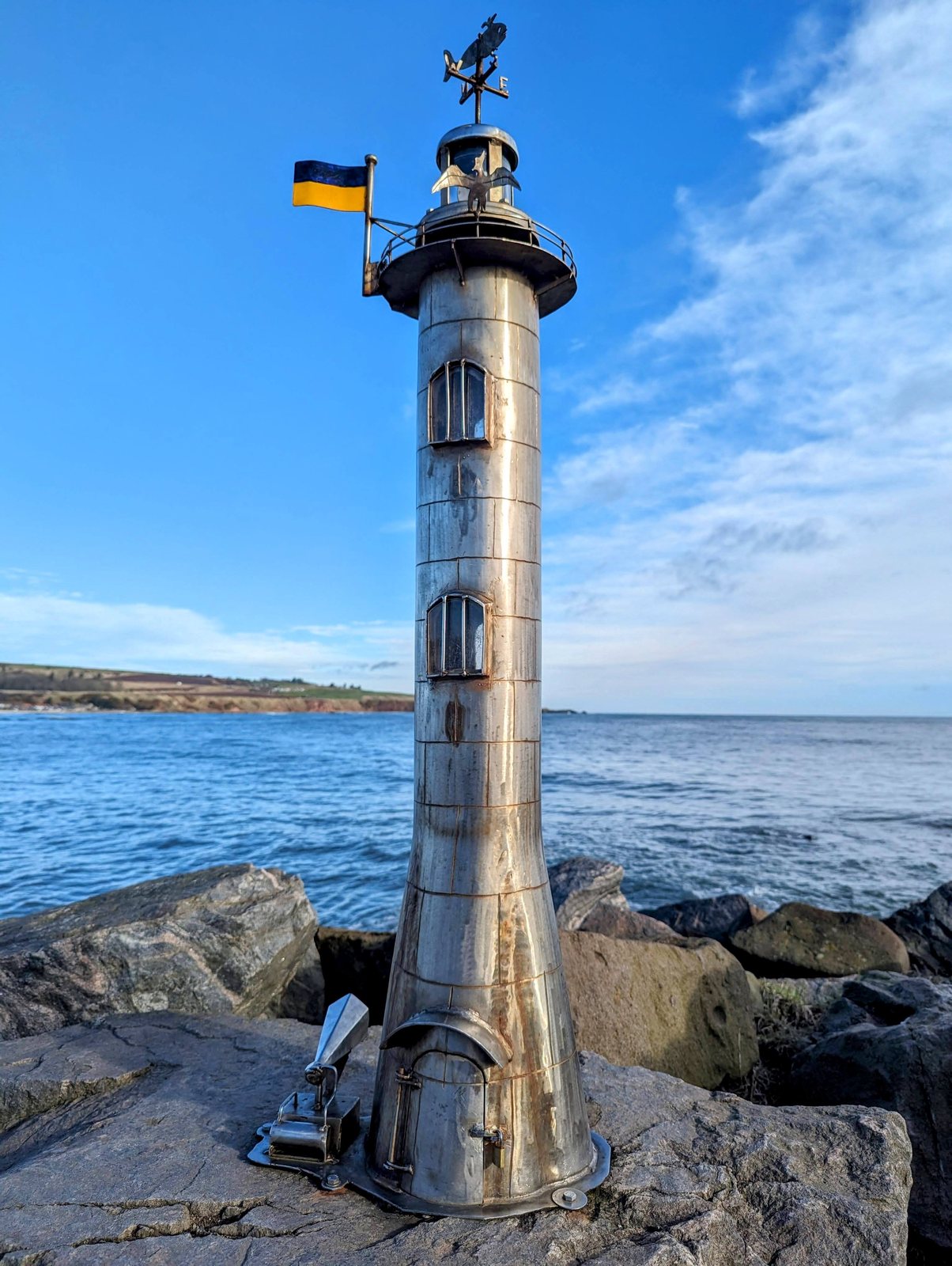 A metal lighthouse sculpture flying a small Ukrainian flag, Stonehaven.