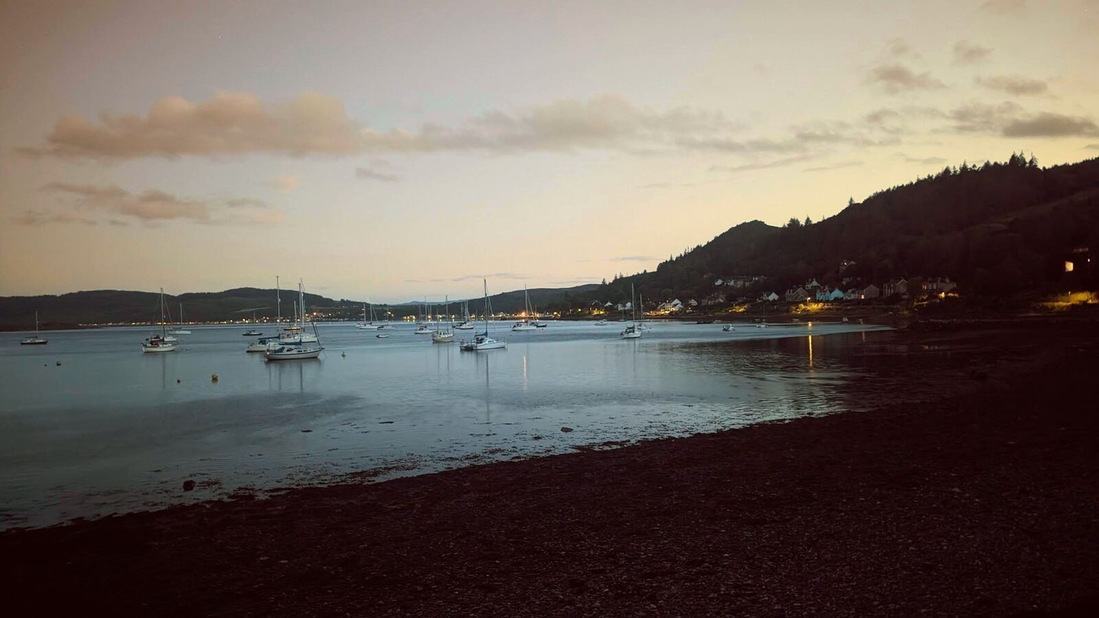 Moored yachts silhouetted on the Kyles of Bute at dusk, village lights beginning to show.